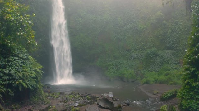 Amazing Nungnung waterfall, Falling water hitting water surface, some huge rocks seeable in front of frame. Lush green leafes is moving from the wind, Bali, Indonesia