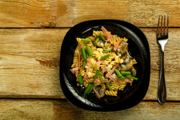 Black plate with pasta with ham and mushrooms in a creamy sauce on the table with a fork.