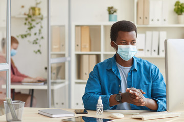 Portrait of young African-American man wearing face mask and sanitizing hands while working in...