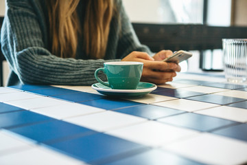 A girl drinks hot tea or coffee in a cafe and uses a cell phone or mobile phone.