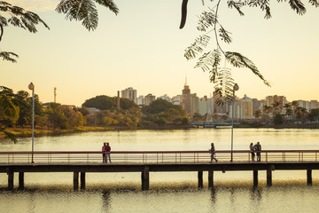 Wooden bridge at the municipal reservoir park in São José do Rio Preto/SP, silhouetted people walking across during a golden sunset, with city buildings in the background