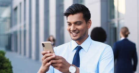 Handsome Hindu young businessman in tie using smartphone and texting message at street. Man from India in business style tapping and scrolling on mobile phone. Outdoors. Close up.