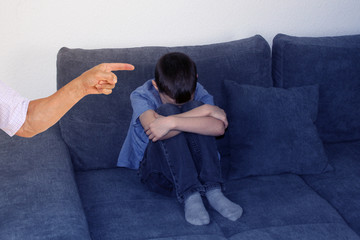 sad child, boy sitting on a gray sofa in the room, clasping his knees with his hands and covering his face, the hand of the parent accusing finger, concept of bad mood and education