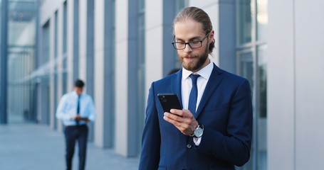 Fototapeta premium Handsome young Caucasian man in glasses standing outdoor and texting message on mobile phone at business center. Male tapping and scrolling on smartphone at street. Businessman using telephone.