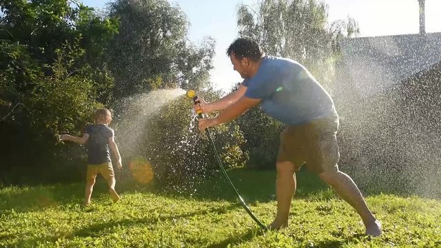 Little boy with his father watering each other with a garden hose in sunny backyard. Preschooler child having fun with spray of water. Summer outdoor activity for family with kids.