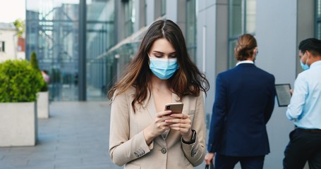 Caucasian young woman in medical mask walking the street and tapping on scrolling on smartphone. Pretty businesswoman using mobile phone and texting message outdoor. Covid-19 concept.
