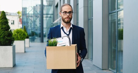 Sad Caucasian man in glasses, white-collar worker walking at street and carrying box with stuff. Fired male. Upset Office manager dismissed from work. Economic crisis of unemployment. Firing from job