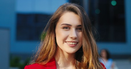 Close up of face of young beautiful pretty woman with long hair smiling cheerfully to camera outdoors. Portrait of Caucasian cheerful happy female with smile in city street. Smiled charming female.