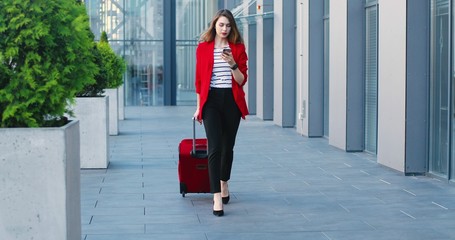 Beautiful young Caucasian woman in red jacket walking and carrying suitcase on wheels, texting on mobile phone at street. Cheerful female tapping on smartphone. Businesswoman at business trip.