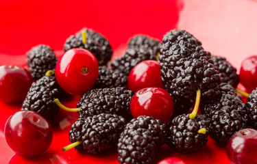 Ripe large black mulberries and red cherries on a red plate, close-up.