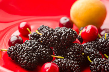 Ripe large black mulberries, peach and red cherries on a red plate, close-up.