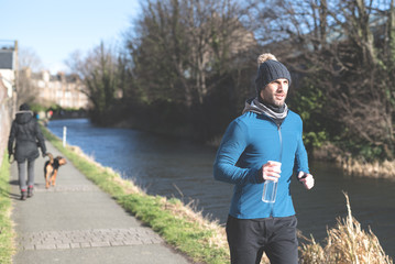 A man wearing a fitted hooded top, leggings and a woolly hat jogs on a footpath along a canal in...