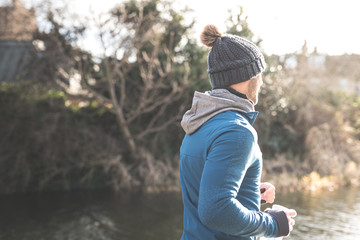 A man wearing a fitted hooded top and a woolly hat jogs on a footpath along a canal with trees on the background in the city of Edinburgh, Scotland, United Kingdom, with the sun on his face