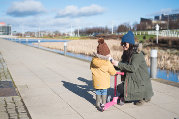 An Asian mother wearing a wooly hat helps her daughter get ready to ride her scooter near a canal in a cold sunny day in the city of Edinburgh, Scotland, United Kingdom