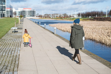 A mother walks near a canal while her daughter pushes a scooter, both wearing woolly hats in a cold sunny day in the city of Edinburgh, Scotland, United Kingdom, where buildings can be seen behind