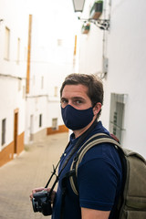 Photo of a young and attractive traveller visiting a city isolated with a photo camera and a backpack. He is wearing a face mask. Holidays with social distance