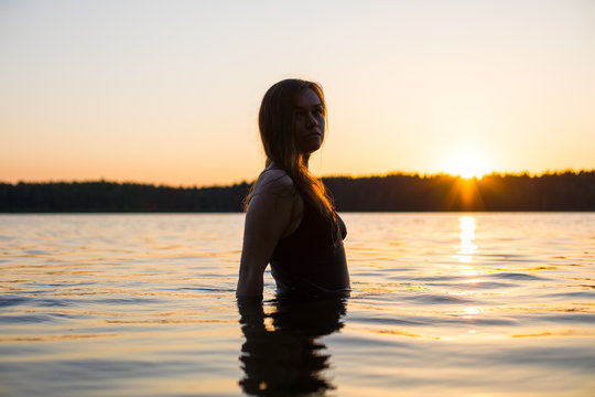 Beautiful Russian girl in a long black swimsuit swims outside the city on the lake in the rays of sunset or dawn. Splashing water on the background of the sun. Happy girl.