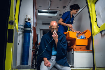 Man breathe through oxygen mask, young nurse packs equipment in medical bag in the ambulance car.