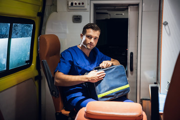 A young man in a medical uniform sitting in an ambulance, holding his medical kit.