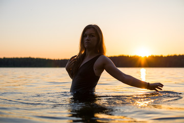 Beautiful Russian girl in a long black swimsuit swims outside the city on the lake in the rays of...