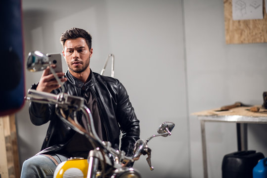 A motorcycle rider in a leather jacket with a smartphone in hand poses for social media and a video blog in a garage
