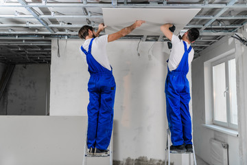 Two handymen are making a plasterboard ceiling. They use a drill and ladders - obrazy, fototapety, plakaty
