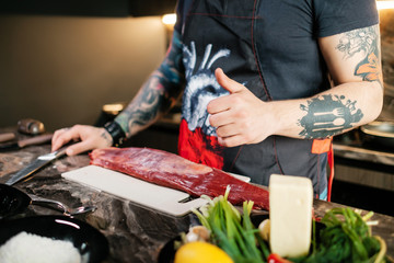 A tattooed chef showing a thumb up before starting to cook a juicy steak at home. Beef on a cutting board with a knife and vegetables. Concept of correct cooking of meat by tattoed butcher.