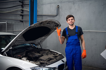 Repairman rocker shows a gesture with his fingers. He is standing next to a white car