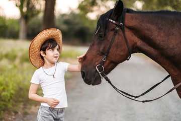 Obraz premium Cute little boy in a cowboy hat stroking a beautiful horse outdoors