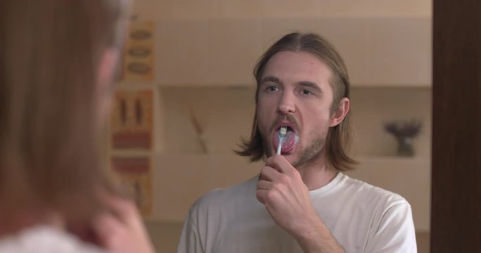 Young man looks at himself in the mirror and brushing tongue with a toothbrush