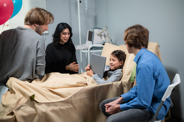 Obraz premium A girl talking to a family member, who couldn't come to visit, over the internet on her tablet, she is staying in a hospital bed, the rest of a family is surrounding her.