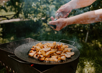 juicy fried potatoes with a crust cooked in a large black pan on the grill. blurred background of green grass and trees. smoke is in the hot summer air.