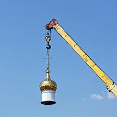 Fototapeta na wymiar The dome of a Christian temple hangs on the hook of a crane. Building or dismantling a church against a blue sky