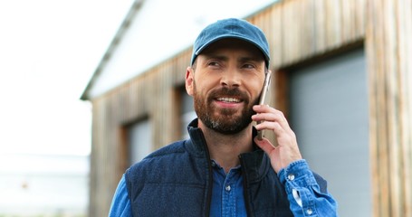 Close up of handsome caucasian man with beard and in hat standing outdoor and talking on mobile phone at wooden shed. male farmer speaking on cellphone. Telephone call. Conversation.