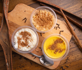 Assortment of traditional Brazilian Festa Junina desserts – rice pudding, peanut candy paçoca, and sweet corn pudding, served in plastic cups with cinnamon, rustic wooden background.