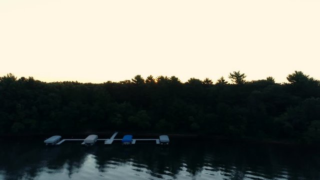 Aerial shot of a pier on the shore of a country lake at sunrise. The drone strafes the pier with boats as the orange rays of the rising sun appear and hide just behind the treetops.