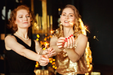 Obraz na płótnie Canvas Selective focus. Sparklers in the hands of two beautiful bokeh woman Christmas garlands and gifts. Gold Christmas decoration. Happy New Year.