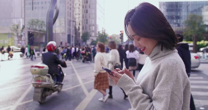 Side view slow motion of happy young asian woman using mobile phone browsing social medial in the urban street with people walking in the background  at Chengdu Sichuan China 