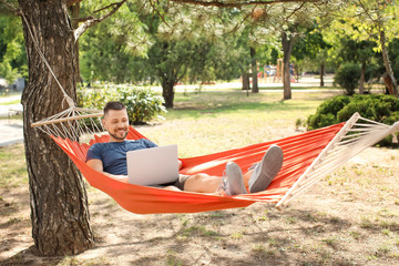 Handsome man with laptop relaxing in hammock outdoors