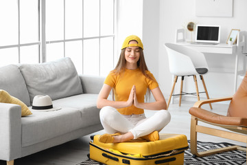 Young woman meditating while sitting on suitcase at home. Travel concept - obrazy, fototapety, plakaty
