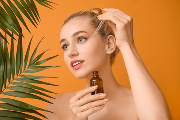 Beautiful young woman with cosmetic oil and tropical leaves on color background