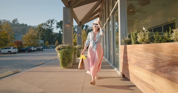 Attractive stylish woman in pink dress with paper bags walking by shopping mall. 4K slow motion girl shops at retail stores in the outdoor shopping village at sunny summer day. California, USA