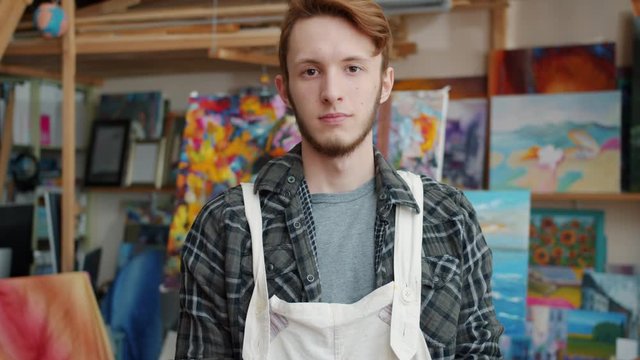 Slow motion portrait of handsome young man art student in modern painting studio standing alone wearing apron and looking at camera with serious face
