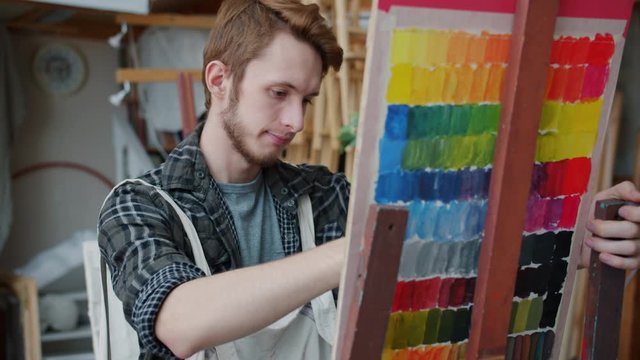 Portrait of serious guy wearing apron painting in art studio concentrated on creative activity working alone in classroom full of beautiful colorful pictures.
