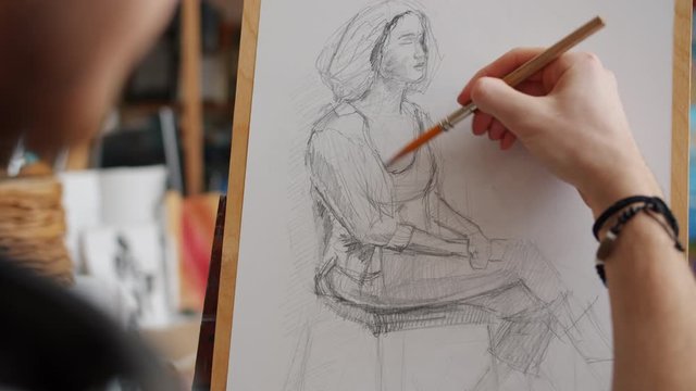 Close-up of man's hand sketching portrait of woman during lesson in art school using pencil on easel working indoors in studio