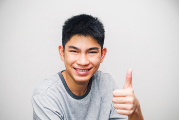 Portrait of a young Asian man smiling with his thumbs up, expressing excellence, or showing successful gestures. In positive human facial expressions and emotions on the white wall background.