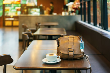 Mini alcohol gel bottle to kill Corona Virus(Covid-19) hang on a brown leather shoulder bag with hot coffee on table in cafe.New normal lifestyle.Health care concept.Selective focus on alcohol gel