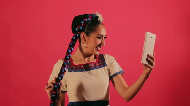Joven mujer mexicana latina en traje regional folclórico utiliza una tableta para tomarse fotos selfie en fondo rosa