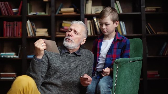 Stylish bearded gray-haired grandfather with his lovely grandson enjoy memories watching family photo album sitting on armchair and floor at home in cozy room with an authentic aristocratic interior.