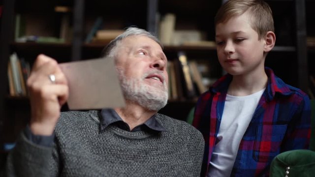 Handsome bearded gray-haired grandfather with his cute grandson having fun looking at an old photo album, enjoying memories watching family photo album at home in cozy room background on bookshelves.